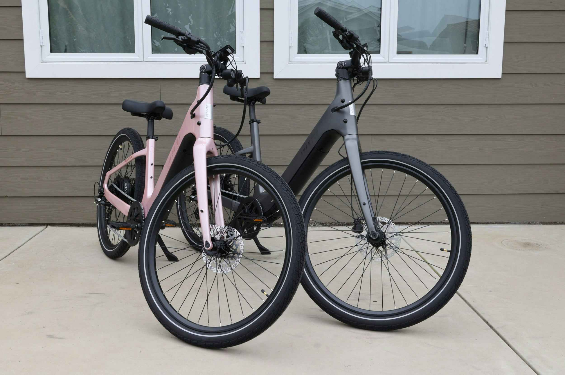 Two modern bikes, including a pink Aventon Pace 5 REC Step-Through electric bike and a black comfort cruiser, are parked on concrete in front of a brown house with white-trimmed windows.