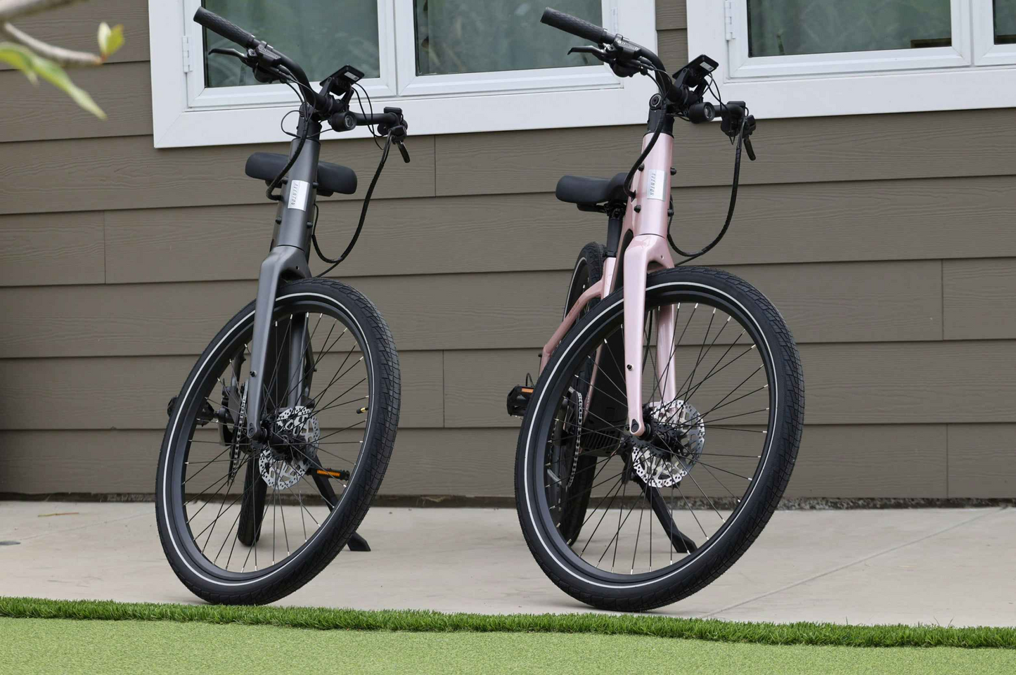 A gray Aventon Pace 5 REC Step-Through and a pink comfort cruiser step-through electric bike are parked upright on a paved surface in front of a house with brown siding and white-framed windows.