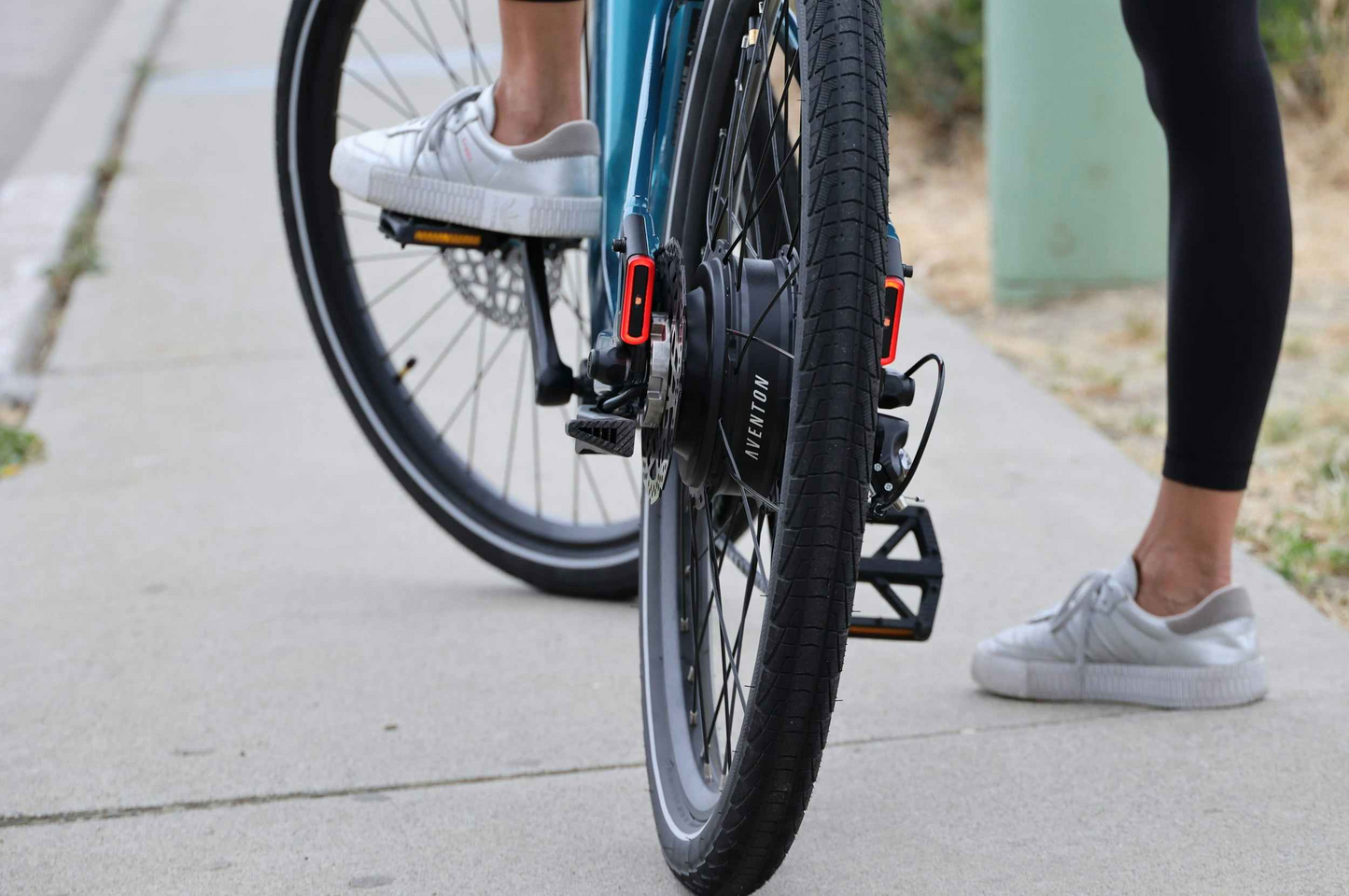 Close-up of two people in white sneakers standing next to their Aventon Pace 5 REC Step-Through electric bikes by Aventon, highlighting the step-through frames and wheels on a sidewalk.