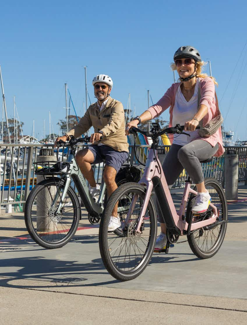 Two adults in helmets ride Aventon Pace 5 REC Step-Through comfort cruiser bikes on a paved path by a marina with boats docked in the background.
