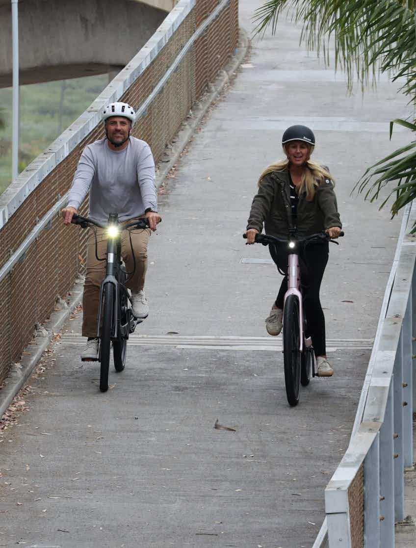Two people in helmets ride Aventon Pace 5 REC Step-Through electric bikes by Aventon side by side on a paved path bordered by railings and greenery.