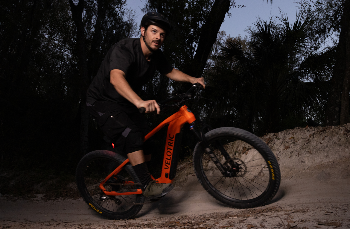 A man in protective gear rides an orange Velotric Summit 1 electric mountain bike on a dirt trail at dusk.