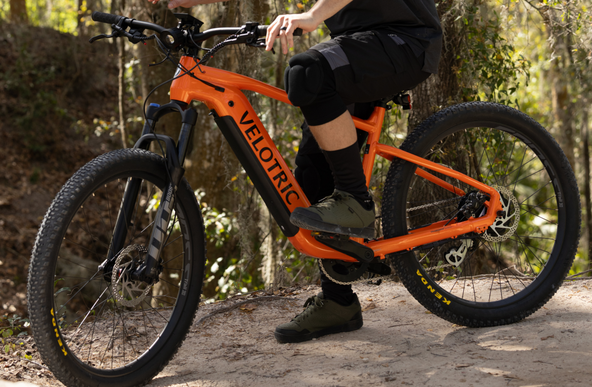 A person in black shorts and boots sitting on an orange Velotric Summit 1 eBike with pedal assist on a forest trail.