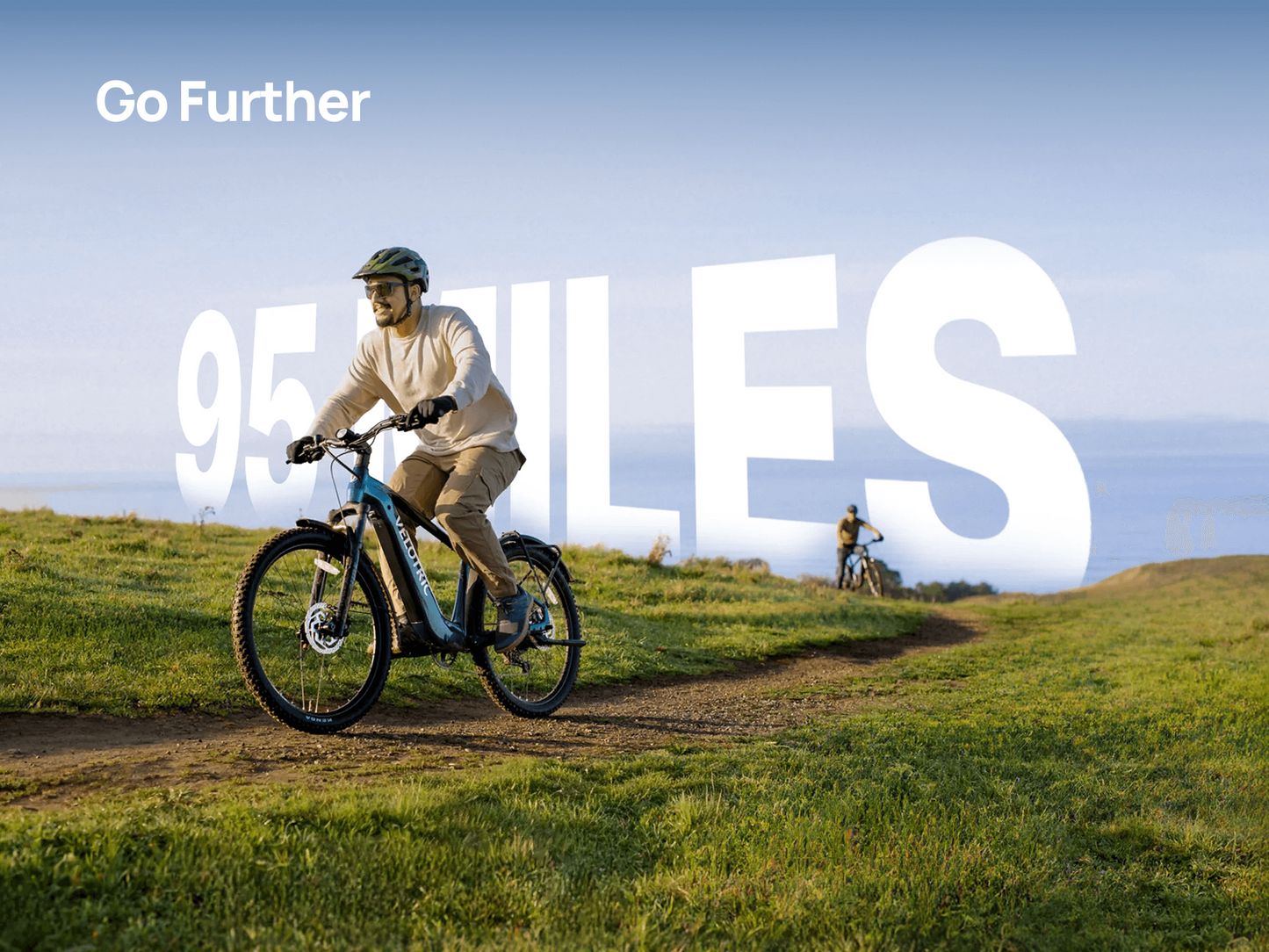 A person rides a Velotric Summit 2 by Velotric on a grassy trail with bold white text "Go Further" and "95 MILES" in the background, as another cyclist appears farther down the path.