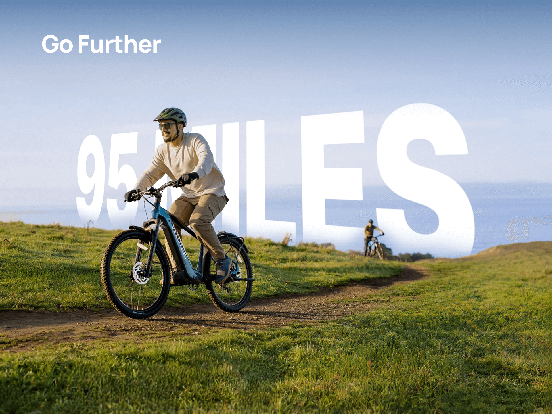 A person rides a Velotric Summit 2 by Velotric on a grassy trail with bold white text "Go Further" and "95 MILES" in the background, as another cyclist appears farther down the path.