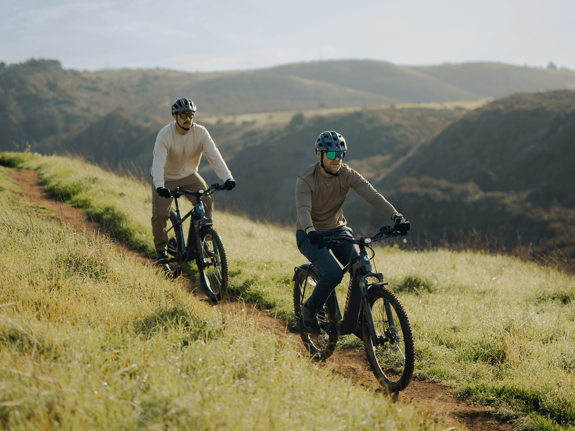 Two people wearing helmets and sunglasses ride Velotric Summit 2 e-bikes by Velotric on a grassy trail, with hills and trees in the background.