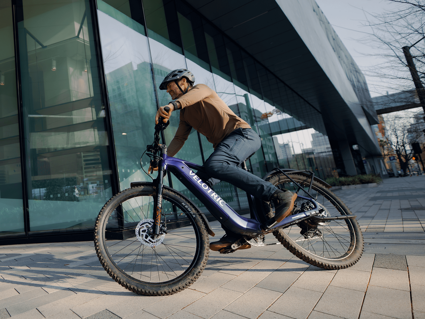 A man in a helmet rides a Velotric Summit 2 eBike by Velotric on a paved urban path beside a modern glass building.