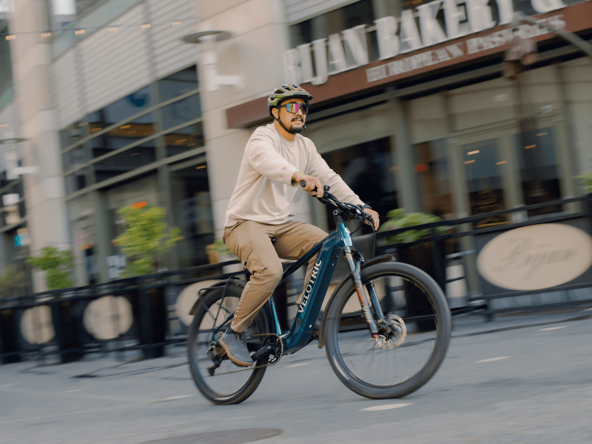 A helmeted rider in sunglasses cruises a city street past a bakery on a Velotric Summit 2 electric bike by Velotric.