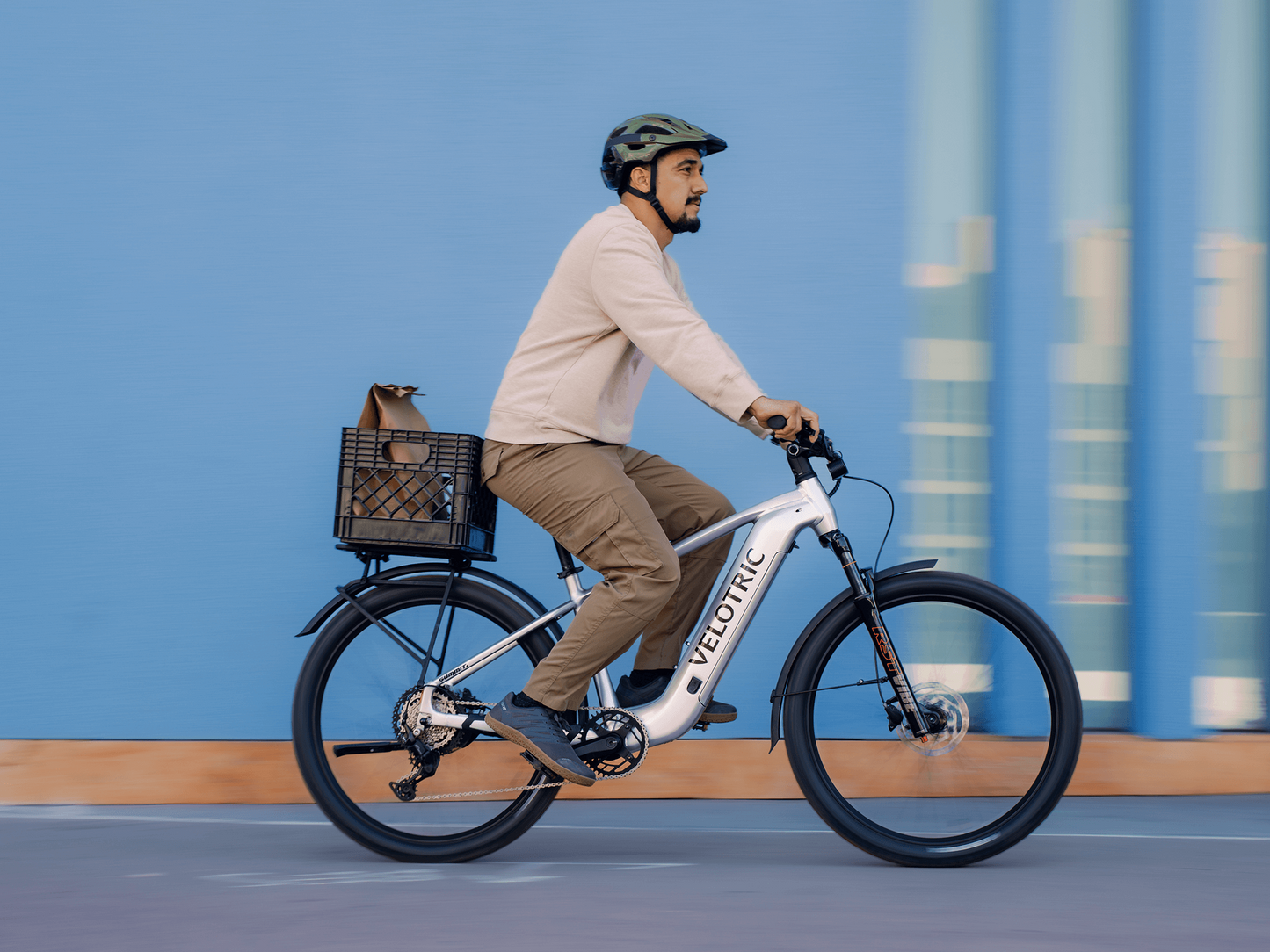 A man in a helmet rides a Velotric Summit 2 hybrid eBike by Velotric, featuring a white frame and rear basket holding a bag, set against a blue wall background.