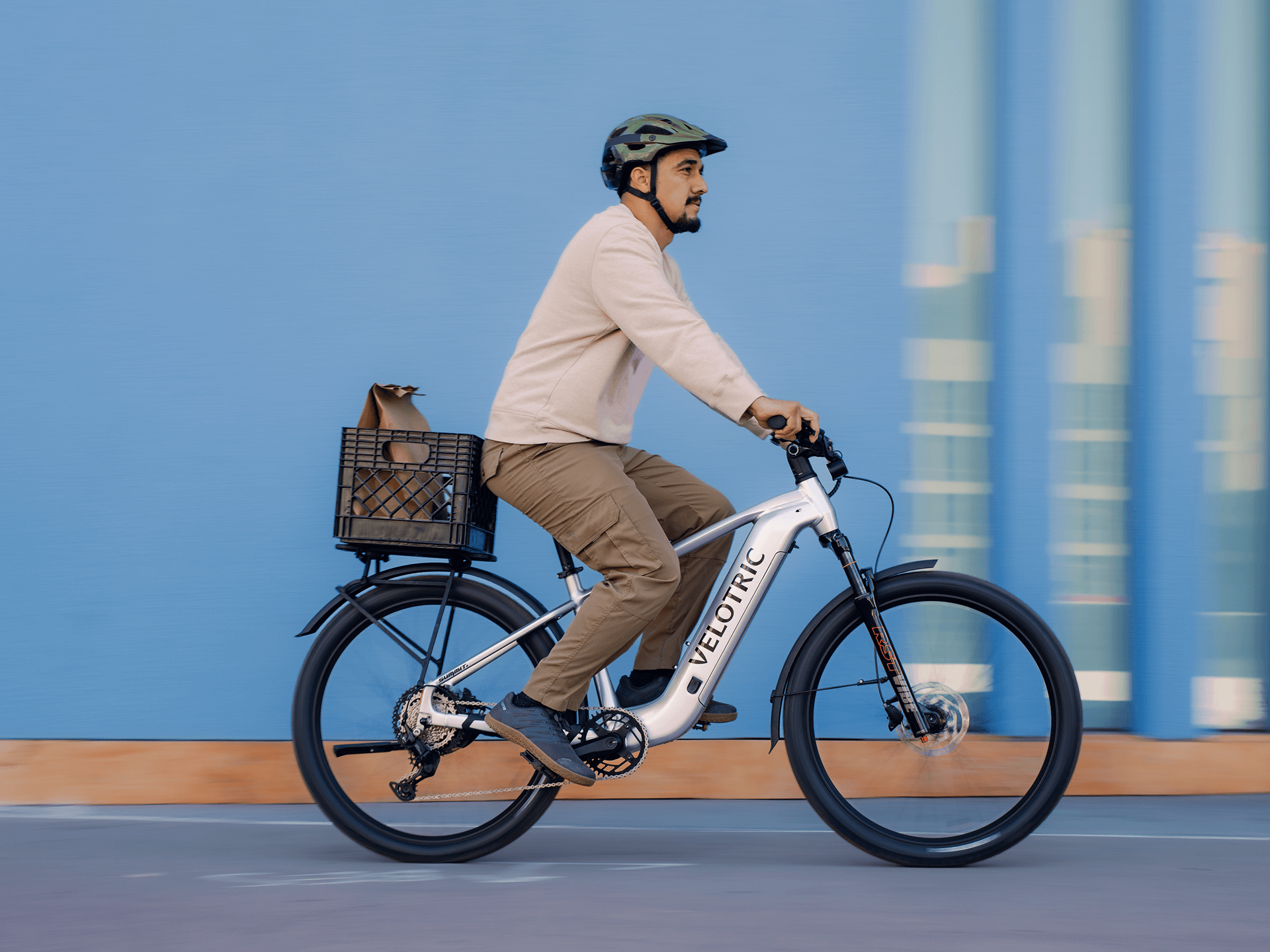 A man in a helmet rides a Velotric Summit 2 hybrid eBike by Velotric, featuring a white frame and rear basket holding a bag, set against a blue wall background.