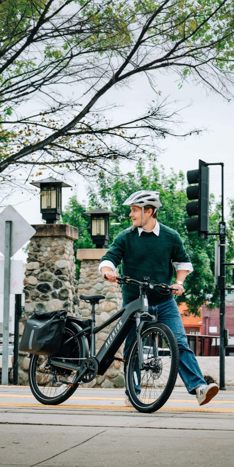 A person wearing a helmet stands beside a black Aventon Level 4 REC electric bike by Aventon on a sidewalk with stone pillars and trees in the background, highlighting its powerful hub motor.