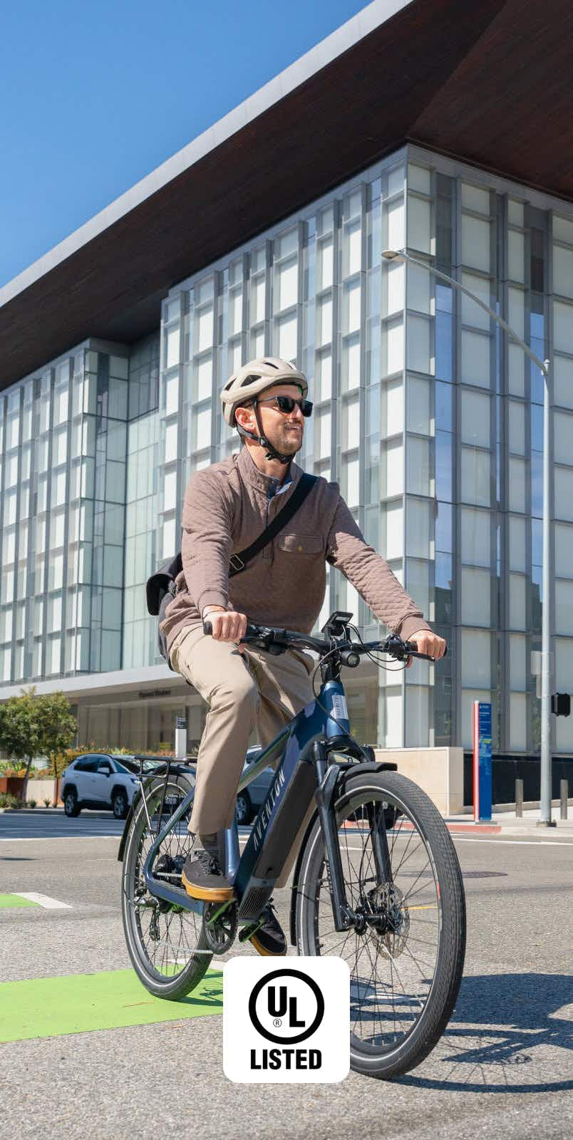 A man in a helmet and sunglasses rides the Aventon Level 4 REC by Aventon, featuring a powerful hub motor, past a modern glass building on a city street. The "UL Listed" certification logo appears at the bottom of the image.