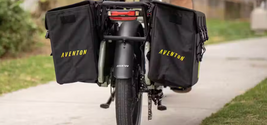 Rear view of a black bicycle with two large water-resistant pannier bags labeled "Aventon Abound" from Tampa Bay eBikes attached to the sides, parked on a sidewalk.