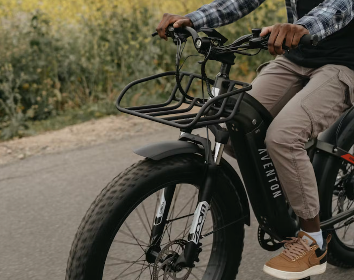 A person in casual attire sitting astride an Aventon electric bike on a paved road, surrounded by autumn foliage, with the Aventon Aventure Front Rack attached.