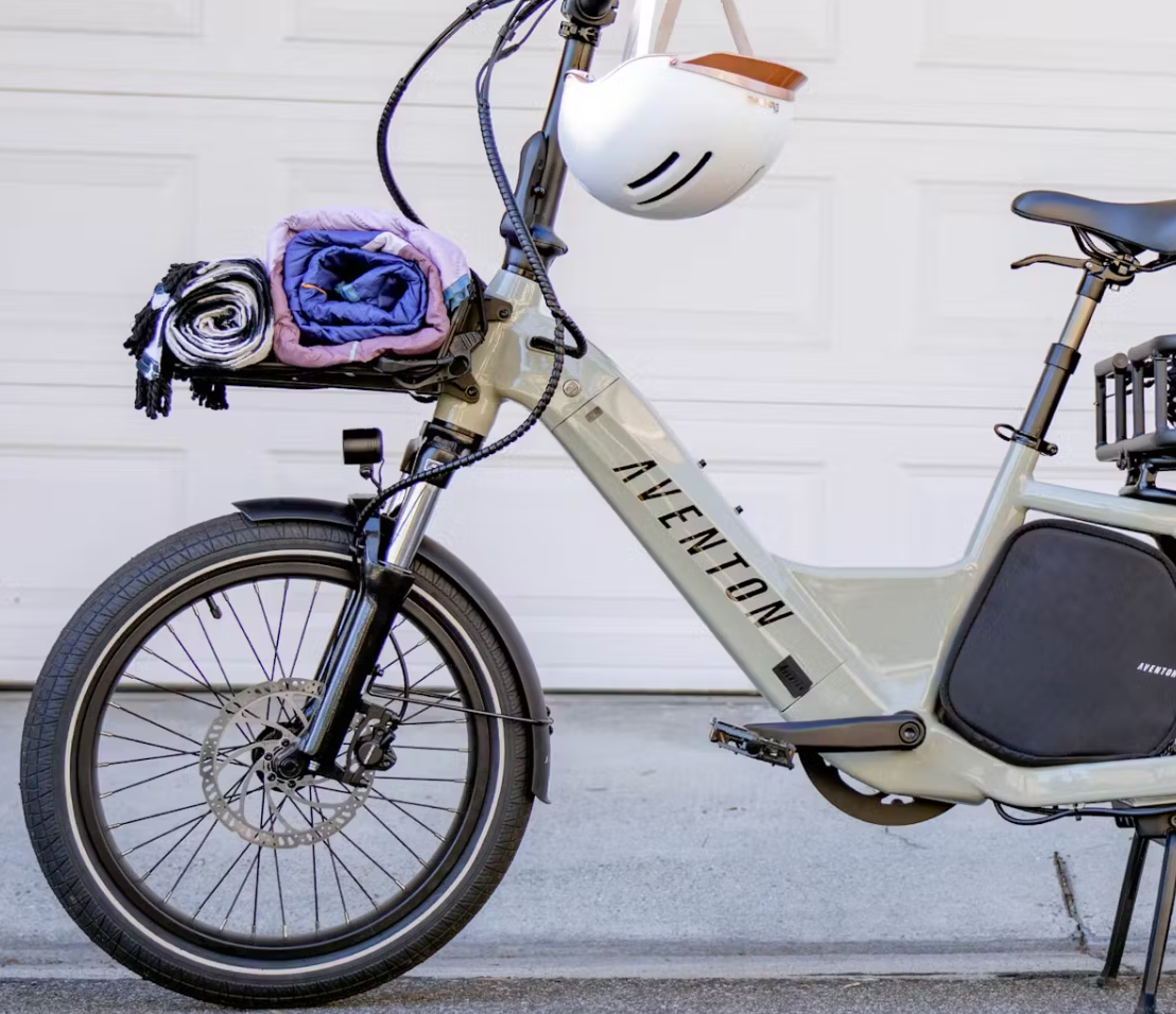Electric bicycle parked against a white garage door, equipped with blankets and a helmet hanging on the handlebars, featuring a Tampa Bay eBikes Aventon Front Utility Rack on the front rack.