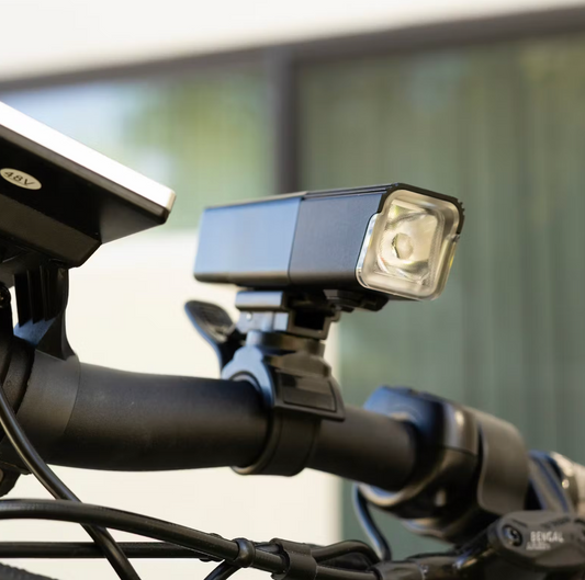 Close-up of a bicycle handlebar showcasing the Tampa Bay eBikes Head Light, a USB rechargeable headlight, with an attached electronic device. The background is blurred.