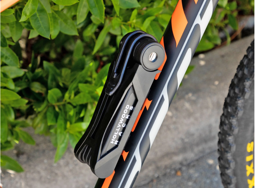 A close-up of a high-security bike lock mounted on the frame of a bike. The lock, branded "Hollywood Racks," boasts "Hollywood Folding" written on it. The background features green foliage and pavement, embodying eBike safety in an urban setting.