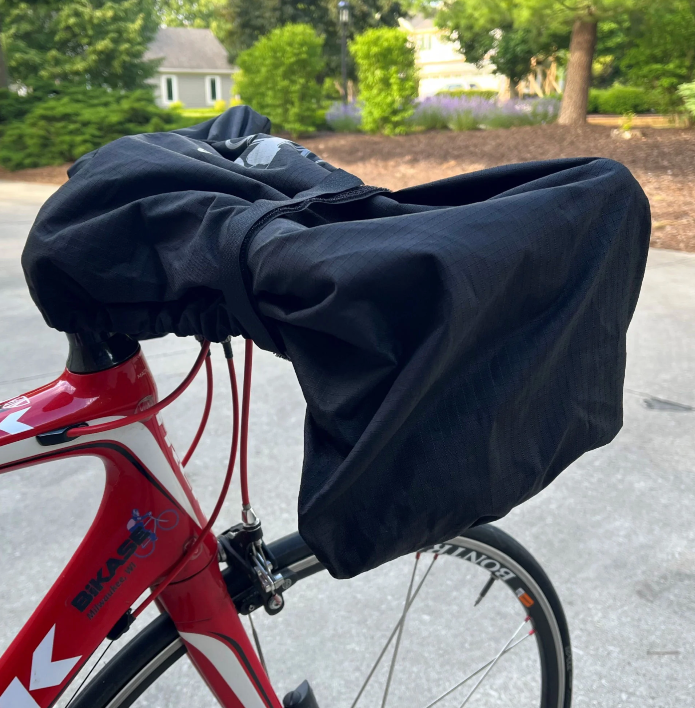 A red bicycle equipped with a Bikase Cockpit Cover, likely protecting it from weather or shielding the handlebars, is parked on a paved area with a garden in the background.