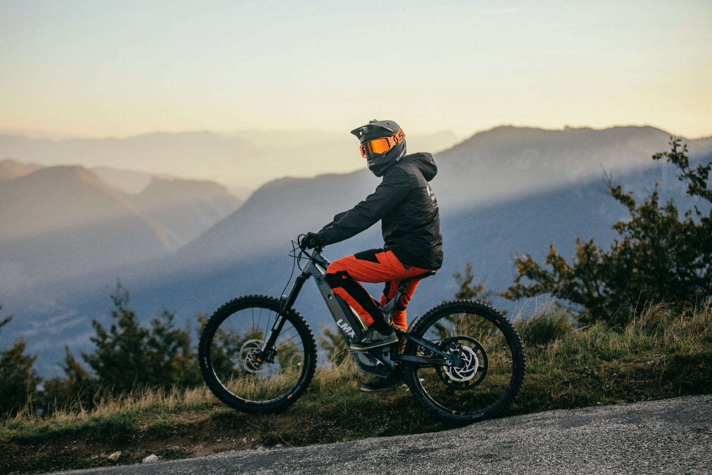 A person in a helmet and protective gear rides an LMX - 64 equipped with a brushless mid-mounted motor on a path with a backdrop of misty mountains at sunrise or sunset.