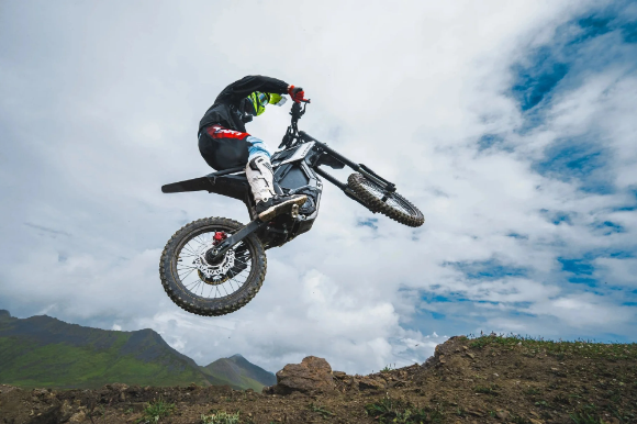 Wearing motocross gear, a rider jumps mid-air over a rocky hill on the Segway Xaber 300 by Segway, demonstrating its impressive off-road performance against a scenic backdrop of mountains and cloudy skies.