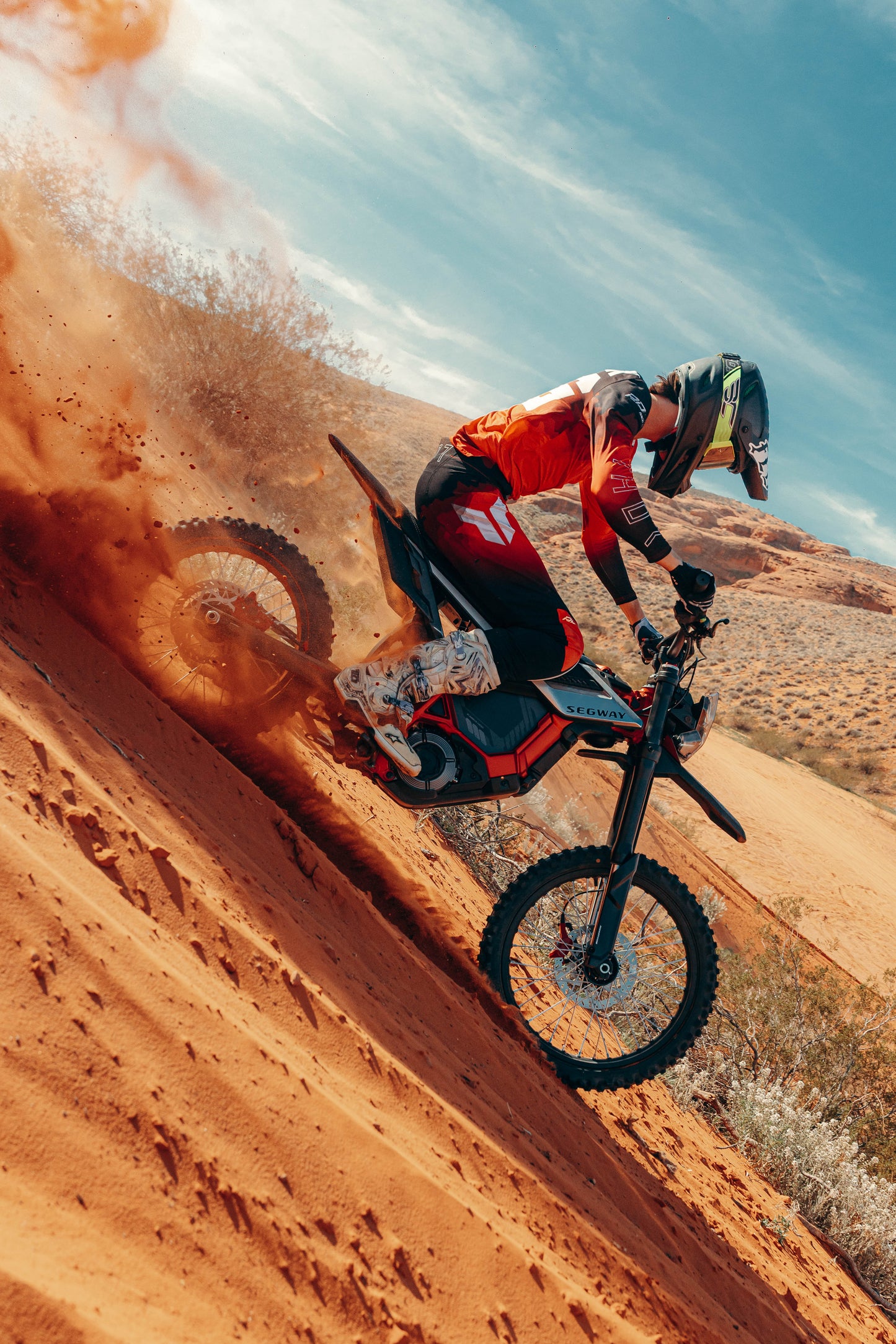 Wearing protective gear, a rider conquers a steep sandy hill on a Segway Xaber 300 by Segway, sending orange sand flying and demonstrating the bike’s impressive off-road capabilities under a partly cloudy sky.