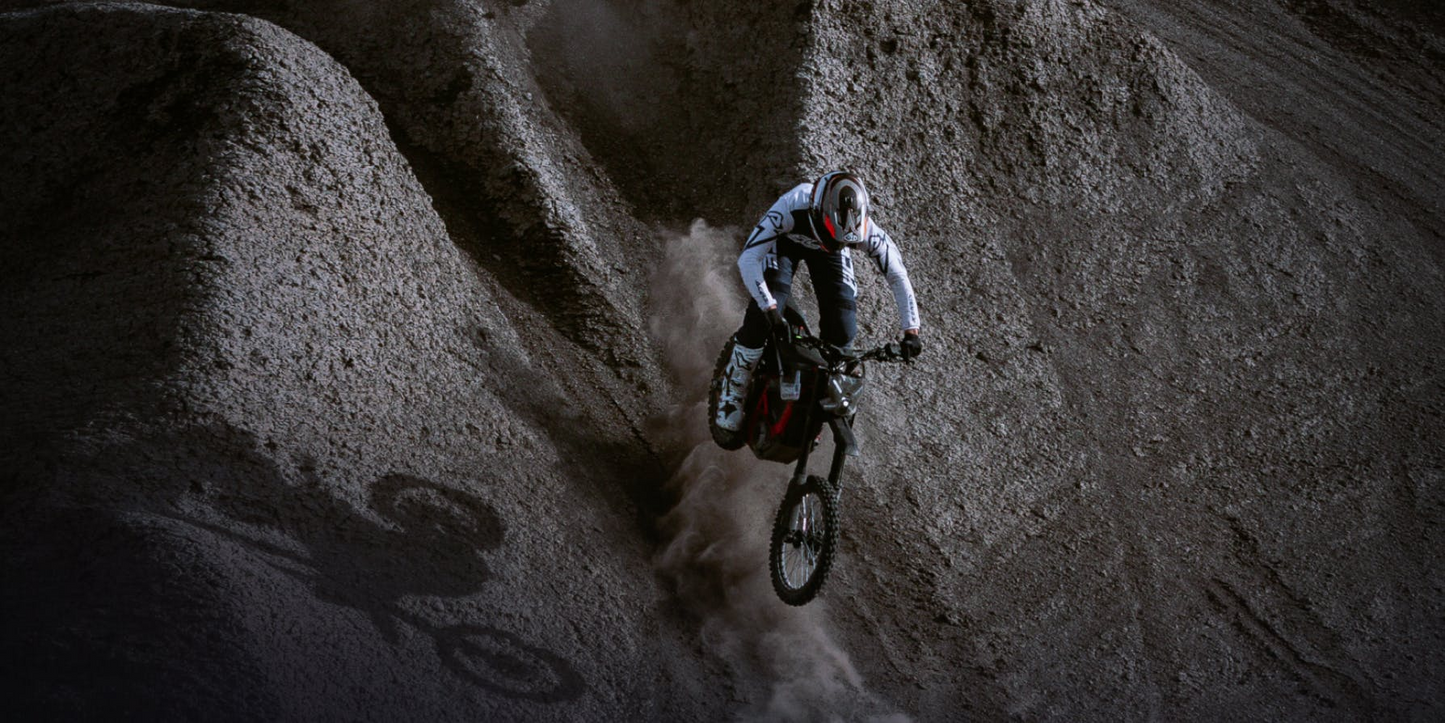 Wearing protective gear, a rider on the Segway Xaber 300 electric dirt bike from Segway descends a steep, dusty slope, casting a shadow on the uneven ground.