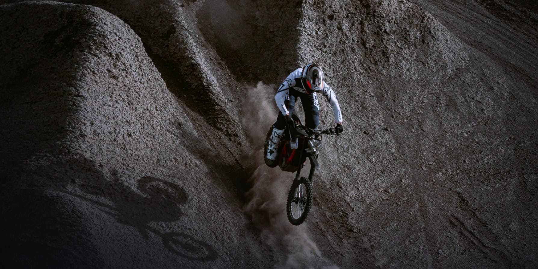 Wearing protective gear, a rider on the Segway Xaber 300 electric dirt bike from Segway descends a steep, dusty slope, casting a shadow on the uneven ground.