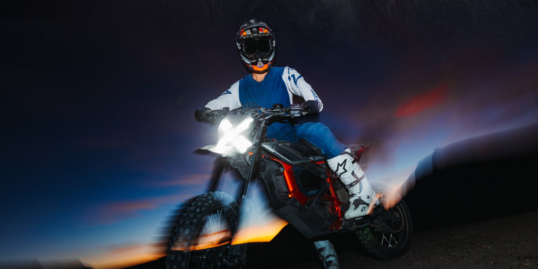 A rider in a helmet and motocross gear rides the Segway Xaber 300 electric dirt bike by Segway at dusk, its headlight shining against a vibrant evening sky.