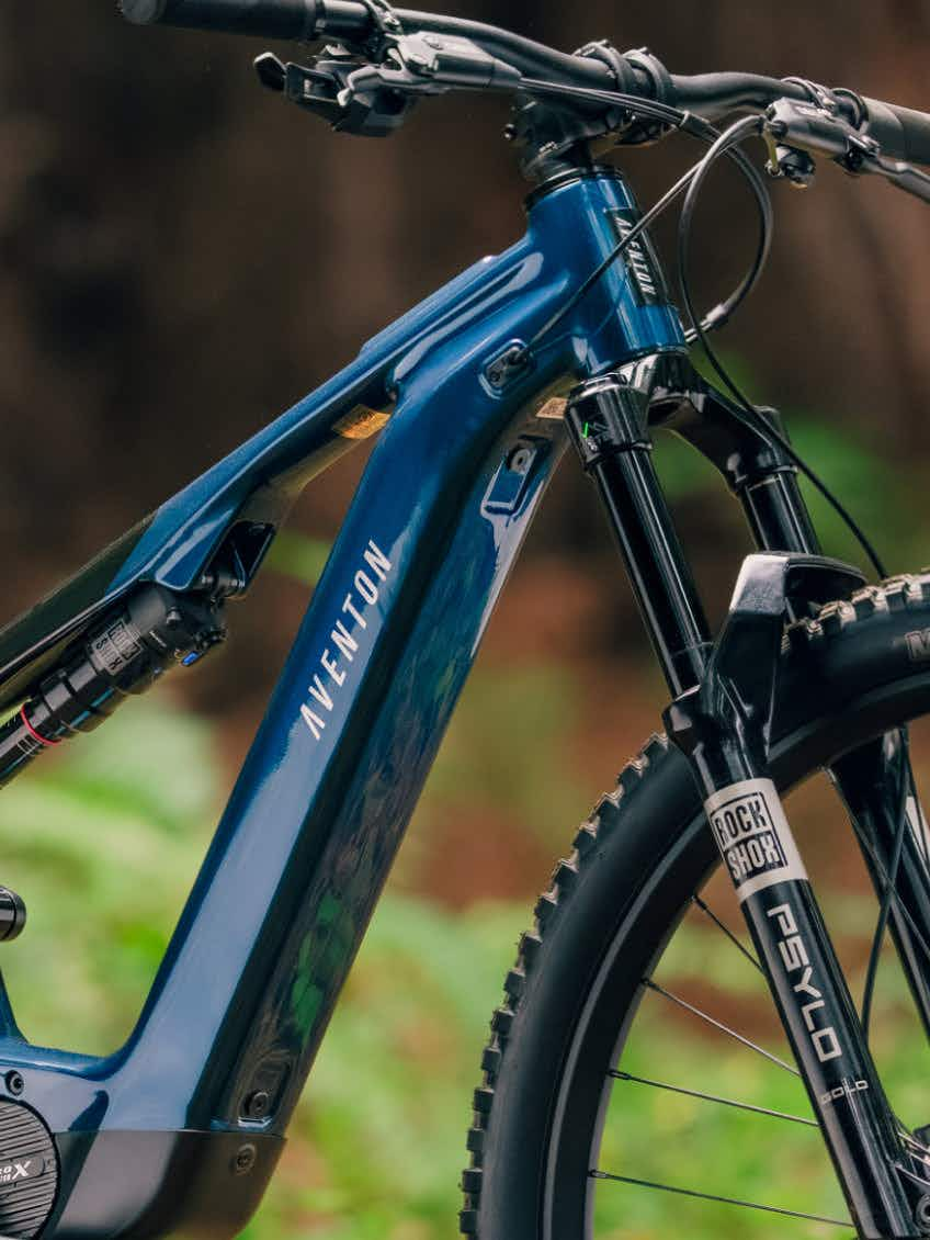 Close-up of the Aventon Current EXP electric bike’s front section, highlighting its blue frame, suspension fork, and front tire with greenery in the background.