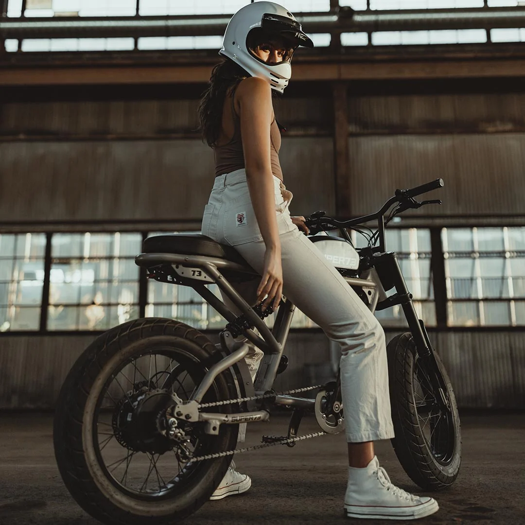 A person wearing a helmet, tank top, and light-colored pants sits on a black Super 73 SUPER73-R Brooklyn SE full suspension electric bike inside an industrial building with large windows.