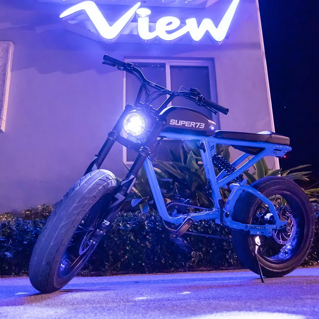 A Super 73 SUPER73-R Brooklyn SE full suspension electric bike, illuminated by its headlight, is parked at night beneath a neon “View” sign, surrounded by plants and a building in the background.