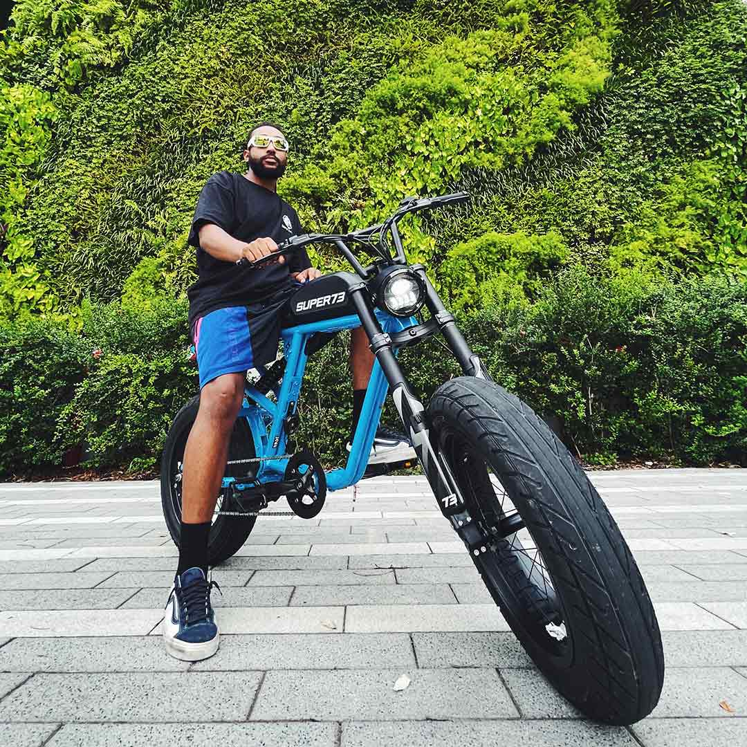 Wearing sunglasses and casual clothes, a person sits atop a blue Super 73 SUPER73-R Brooklyn SE electric bike with full suspension on a paved path, surrounded by greenery and a vertical garden in the background.