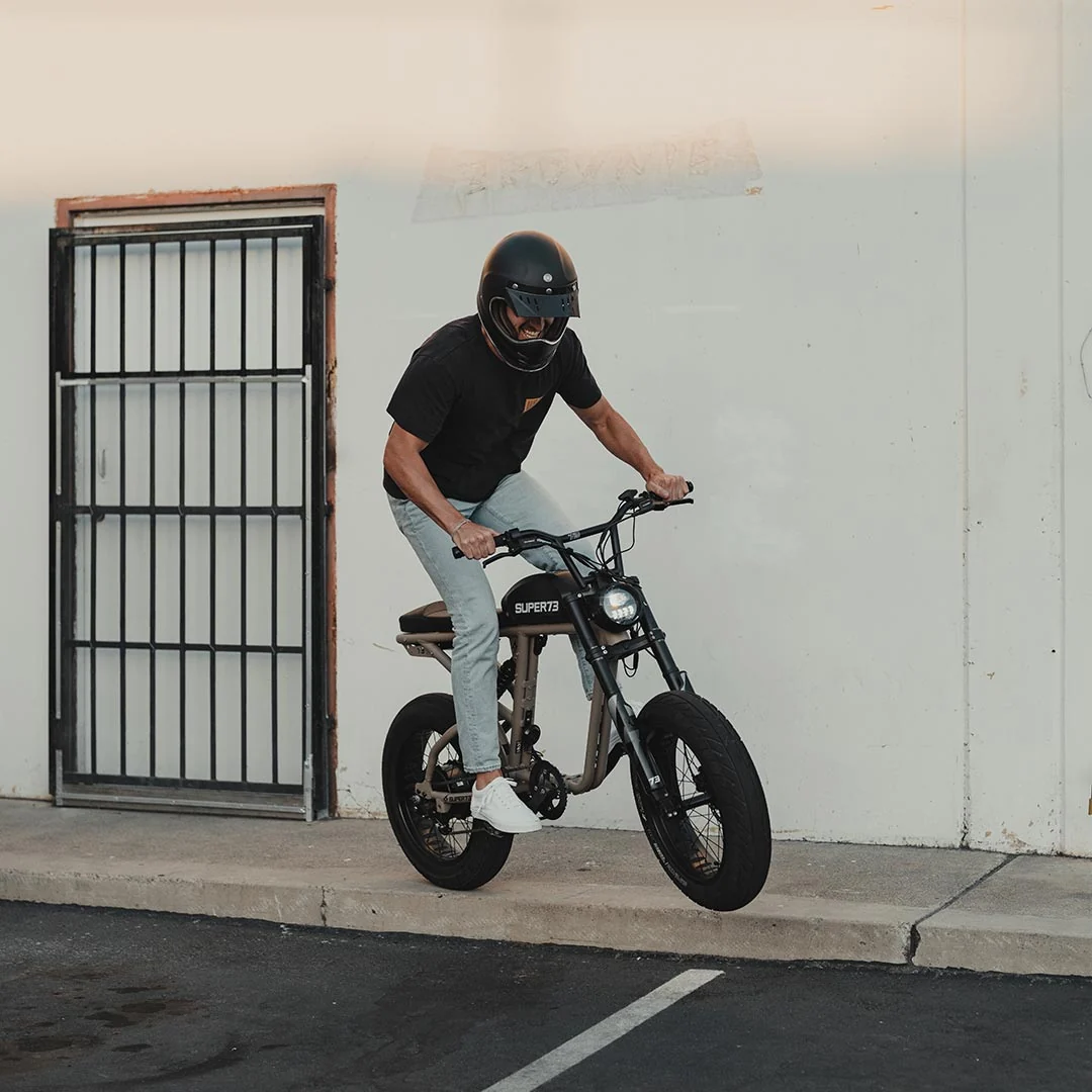 A helmeted individual rides a black Super 73 SUPER73-R Brooklyn SE electric bike, popping a wheelie on a concrete curb beside a metal gate and white wall. The bike’s full suspension from Super 73 provides enhanced control and comfort during this dynamic stunt.