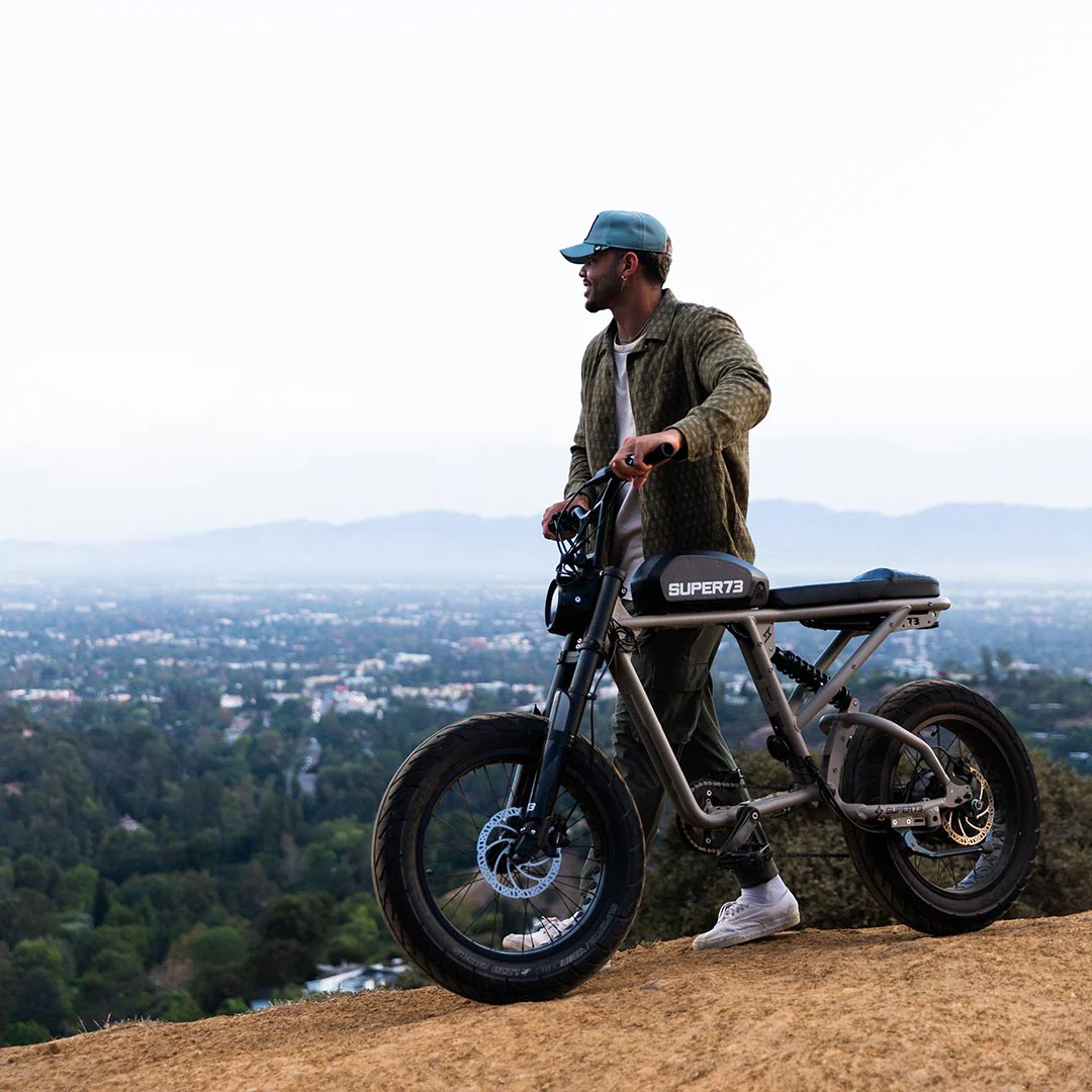 A man stands next to a Super 73 SUPER73-R Brooklyn SE full suspension electric bike on a hilltop, overlooking a cityscape framed by trees, buildings, and mountains in the background.
