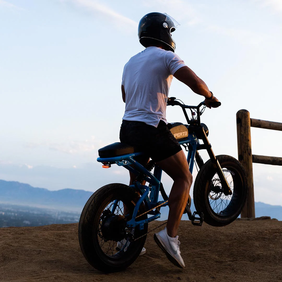 A person wearing a helmet, white shirt, and shorts sits on a Super 73 SUPER73-R Brooklyn SE electric bike, featuring full suspension, on a dirt trail overlooking mountains at sunset.