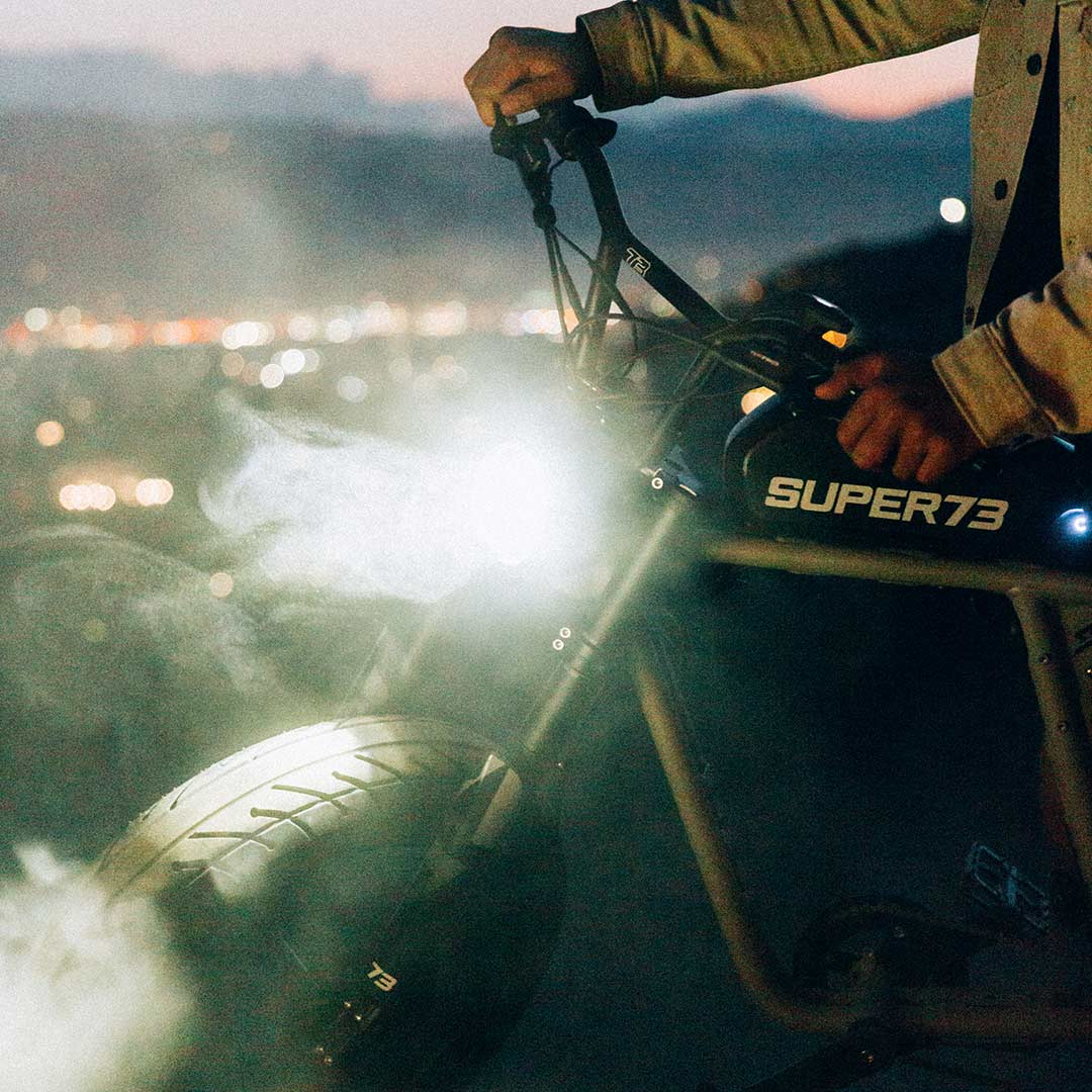 A person grips the handlebars of a Super 73 SUPER73-R Brooklyn SE electric bike at dusk, its headlight illuminating swirling smoke around the front wheel while the full suspension stands out in the fading light.