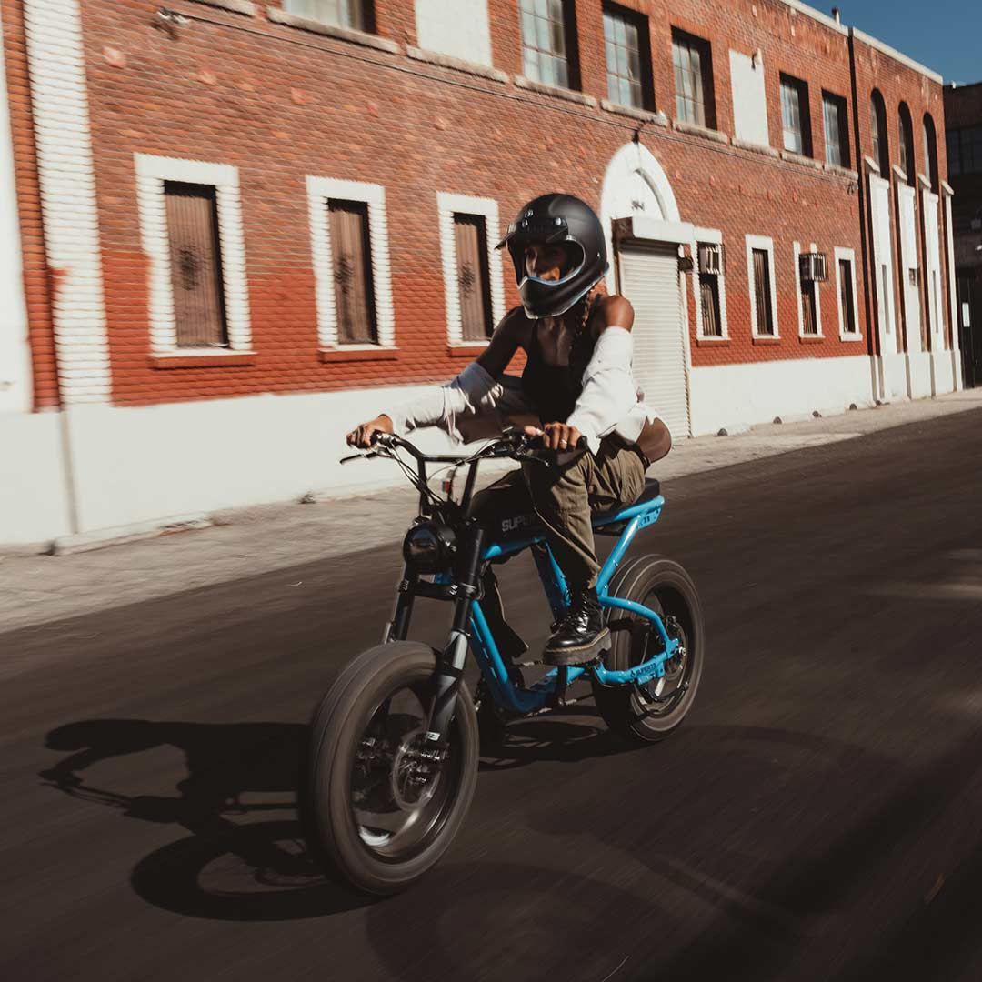 A person wearing a black helmet rides the SUPER73-R Brooklyn SE, a blue full suspension electric bike from Super 73, on a paved street in front of a red brick building.
