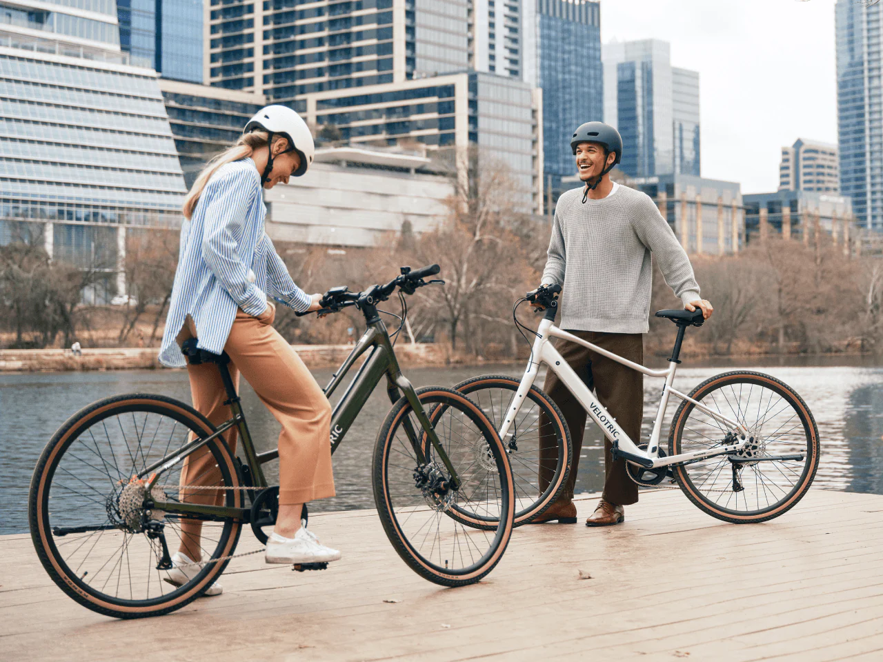 Two people wearing helmets stand by the water with their Velotric Tempo electric bikes from Velotric, a city skyline and trees in the background.