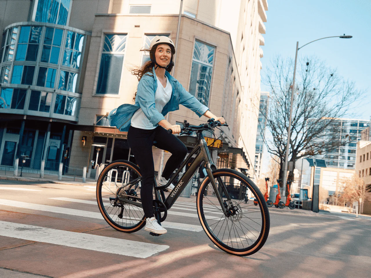 A woman wearing a helmet rides the Velotric Tempo by Velotric across a city street on a sunny day, highlighting the lightweight design and comfort of this electric city bike amid modern buildings and greenery.