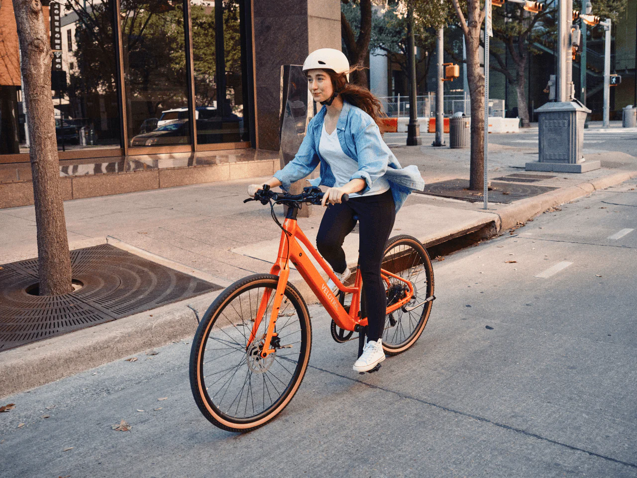 Wearing a helmet and casual clothes, a person rides the Velotric Tempo electric bike by Velotric down a tree-lined street flanked by city buildings.