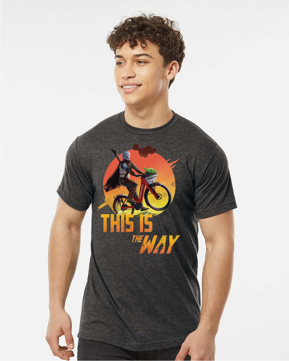 A young man wearing the "This Is The Way" eBike Graphic Tee by Tampa Bay eBikes, which features a colorful character riding a bike along with the text "THIS IS THE WAY" printed on it, stands against a plain background.