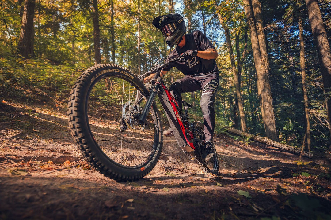 A cyclist in protective gear and helmet rides a mountain bike with pedal assistance down a steep, forested trail on a sunny day. With the advanced brushless mid-mounted motor from LMX - 64 by LMX, navigating the rugged terrain becomes an exhilarating experience.