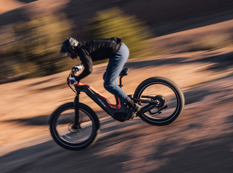 A cyclist in motion on a HeyBike - Hero (Mid-Drive) electric bike, wearing a helmet and black jacket, blurred background indicating speed on a dirt trail.