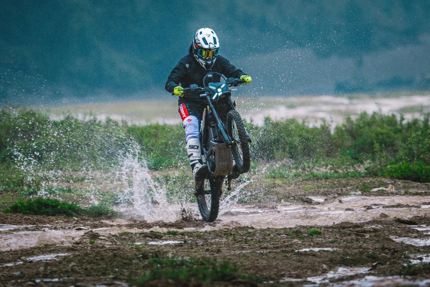 Wearing protective gear, a rider lifts the front wheel of the Segway Xaber 300 electric dirt bike through muddy terrain, as water splashes around, showcasing Segway’s impressive off-road performance.