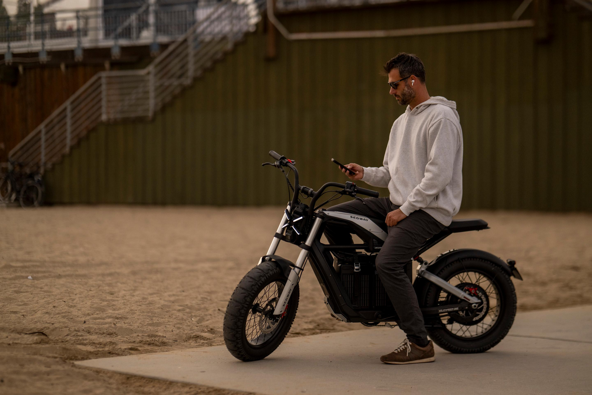 A man in a hoodie sits on his Segway - Xyber, an eBike, on a sandy path. He's absorbed in his phone while stairs and a green wall form the backdrop, all under Tampa Bay's sunlit skies.