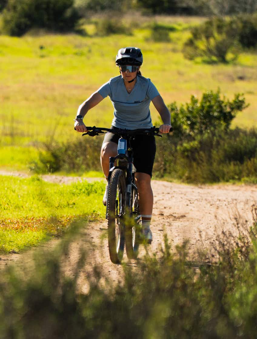 Wearing a helmet and sunglasses, a rider cruises along a sunlit dirt trail on the Aventon Ramblas ADV electric mountain bike by Aventon.