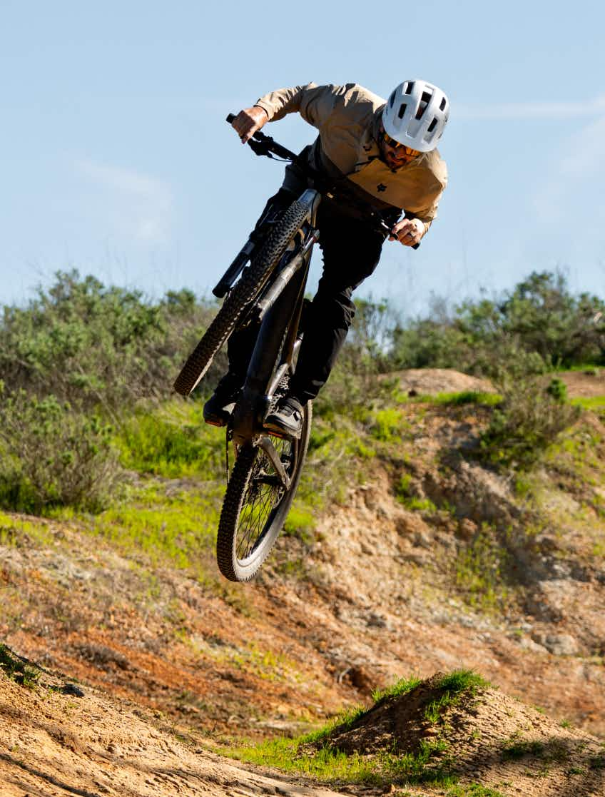 A rider wearing a helmet jumps an Aventon Ramblas ADV by Aventon off a dirt trail, captured mid-air with grass and shrubs in the background.