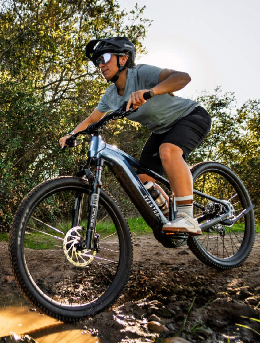 A person in a helmet and sunglasses rides the Aventon Ramblas ADV electric mountain bike by Aventon along a dirt trail through the woods.