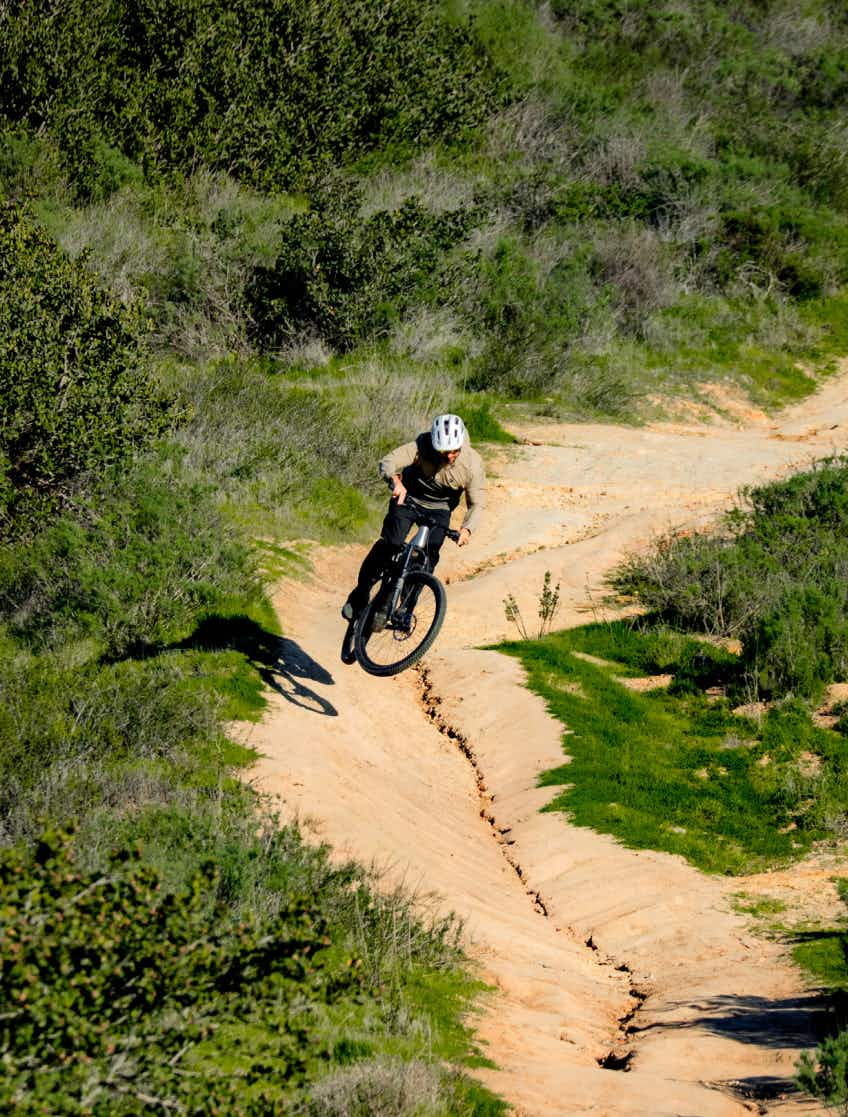A person in a helmet rides the Aventon Ramblas ADV by Aventon, an electric mountain bike, down a dirt trail lined with lush green vegetation.