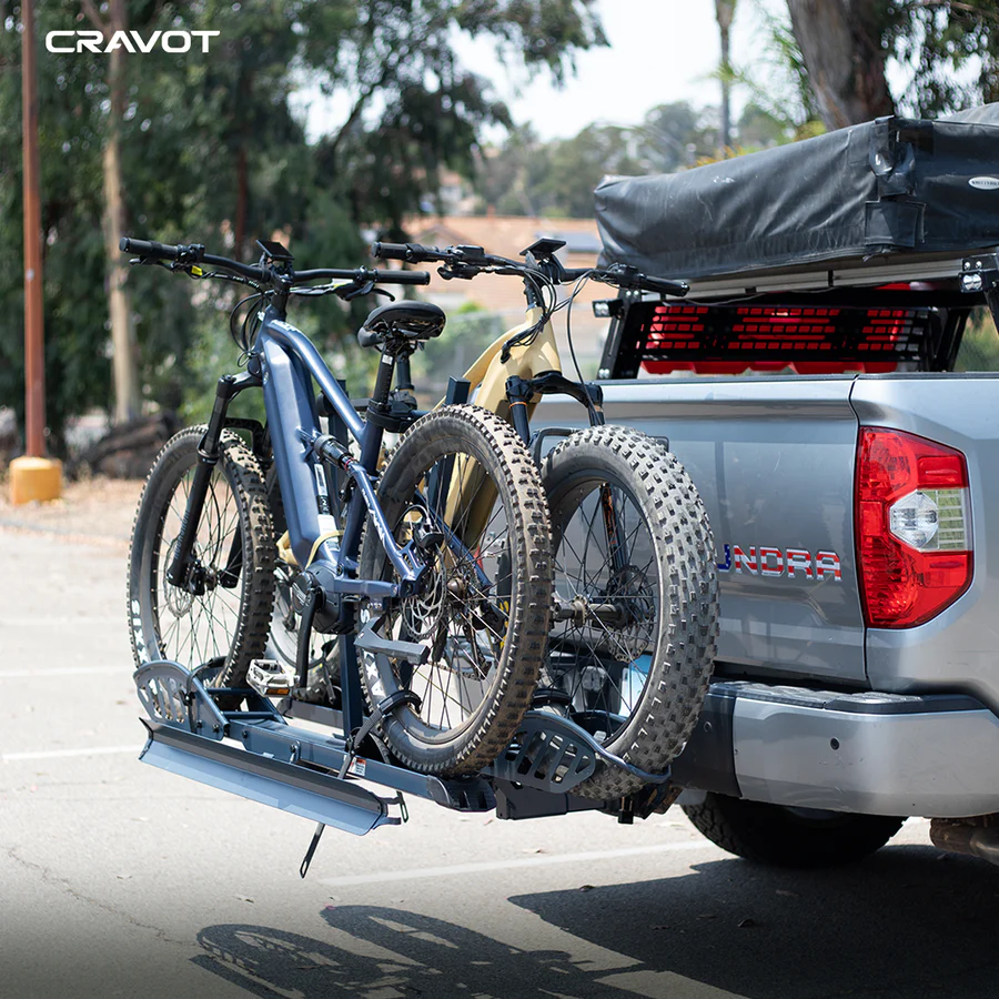Two mountain bikes are mounted on a Cravot CyberRack E2 hitch bike rack attached to the back of a gray pickup truck parked outdoors.